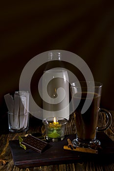 Glass of coffee on a wooden table with a bottle of milk, sugar bags, cologne and spices lit by candles