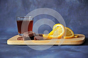 A glass of cognac, chocolate wedges and slices of sliced lemon on a wooden board stand against a dark gray background