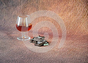 A glass of cognac and a few pieces of chocolate on a brown abstract background. Close up shot