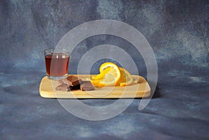 A glass of cognac, pieces of chocolate and lemon wedges lie on a wooden board on a gray background
