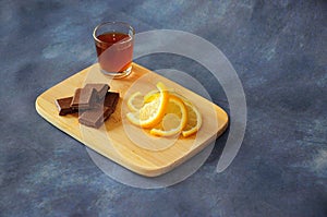A glass of cognac, slices of chocolate and lemon slices stand on a wooden board against a gray background