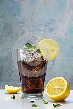 glass of cola or coke with ice cubes, lemon slices and peppermint garnish against a blue vintage wall