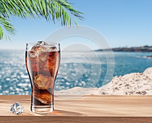 Glass of cola drink with ice cubes on table and blurred sparkling sea at the background