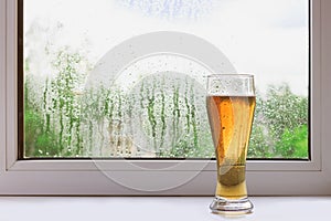 Glass of cold beer on the windowsill on a rainy summer day. The view from the window. Rain drops on glass.