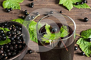 glass of cold black currant juice on wooden table with ripe berries in pottery bowl