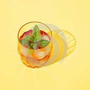 A glass of cold red drink with ice, mint and hers shadows in the sun. Yellow background