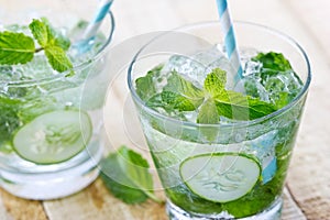 Glass of cold water with fresh mint leaves and cucumber with ice cubes on wooden background