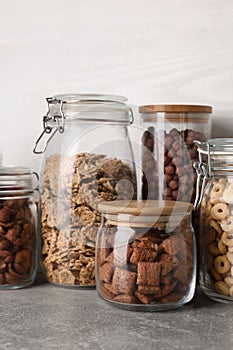 Glass containers with different breakfast cereals on grey countertop near white wooden wall