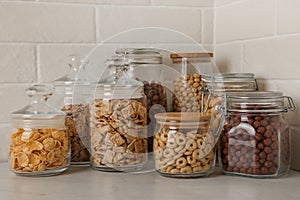 Glass containers with different breakfast cereals on white countertop near brick wall