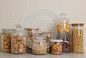 Glass containers with different breakfast cereals on white countertop near brick wall