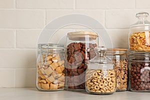 Glass containers with different breakfast cereals on white countertop near brick wall