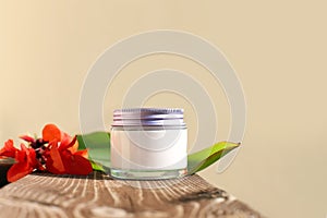 Glass cosmetic jar with white cream and metal screw cap and large tropical green canna leaf an red flower on beige background, on