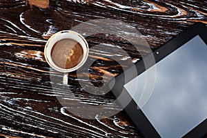 A glass cup of aromatic black coffee, and a black tablet on a dark wooden table