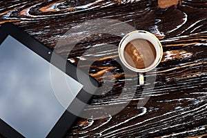 A glass cup of aromatic black coffee, and a black tablet on a dark wooden textured table.