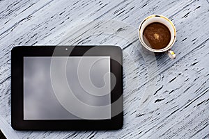A glass cup of aromatic black coffee, and a black tablet on a light wooden textured table