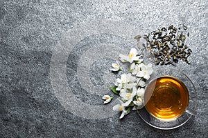 Glass cup of aromatic jasmine tea, dry leaves and fresh flowers on grey table, flat lay. Space for text