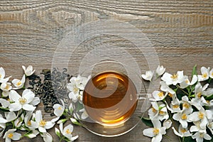 Glass cup of aromatic jasmine tea, dry leaves and fresh flowers on wooden table, flat lay. Space for text