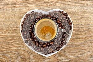 Cup with black coffee on roasted coffee beans in saucer in heart shape on wooden table. Top view