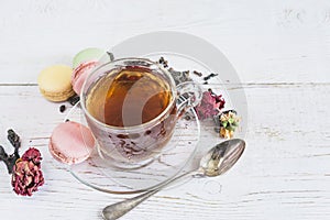 Tea time concept. Glass cup of black tea, french macaroons and dry rose buds on the rustic white paint wooden backgorund