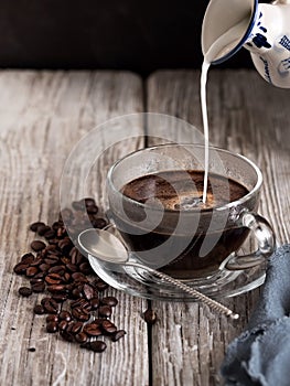 Glass cup with coffee, milk is poured from a vintage milkman. Coffee beans on a wooden background. Close-up