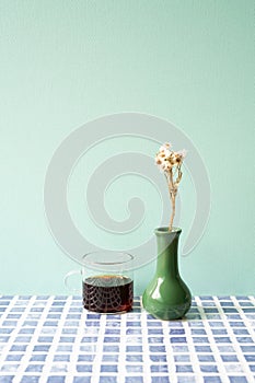 Glass cup of coffee with vase of dry flower on blue tile desk. mint wall background