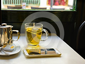 Glass Cup with Freshly Brewed Steaming Hot Green Tea with Pot Honey Smartphone on Table by Window in Cafe