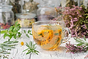 Glass cup of healthy calendula herbal tea, heather, rosemary, chamomile and salvia medicinal herbs and flowers on table.