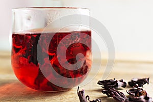 A glass cup with red hibiscus tea stands on a wooden table.