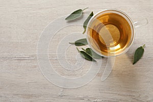 Glass cup of sage tea and green leaves on wooden table, top view. Space for text