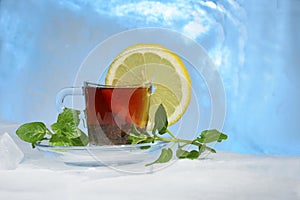 Glass cup of strong black tea on a beautiful blue ice with yellow lemon and green mint.