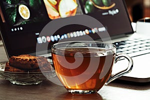 Glass cup with tea and cookies on a office desk with a laptop Tea break from loved job. Work online or offline or in office,