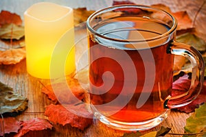 Glass cup of tea and glowing candle on wooden table with autumn leaves.