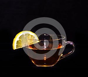 A glass cup of tea with a yellow lemon slice at the border in front of a black background