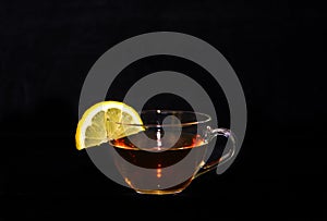 A glass cup of tea with a yellow lemon slice at the border in front of a black background