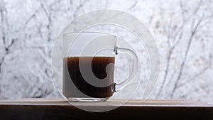 A glass cup on the windowsill into which hot coffee and milk are poured, steam