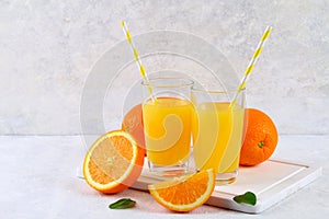 Glass cups and a pitcher of fresh orange juice with slices of orange and yellow tubes on a light gray table.