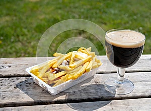 Glass of dark strong belgian beer and portion of fried potato chips with sauce and onion, served on outdoor terrace with green