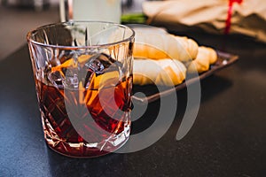 Glass of dark vermouth drink with slice of orange on a bar table outdoors, a popular aperitif, during a summer day with snacks and