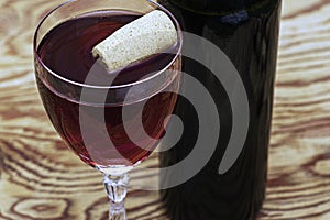 A glass of dry red wine on a wooden table with a cork floating in the glass there is a bottle of wine nearby