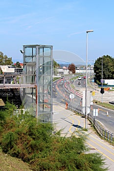 Glass elevator on side of train station surrounded with dense vegetation and street under reconstruction with warning road signs