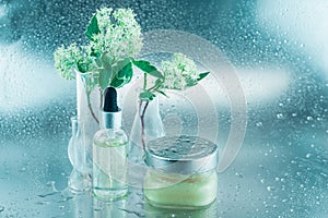 Glass flasks with flowers, a bottle of oil and big jar of cream on a silver background covered with drops. Natural cosmetical