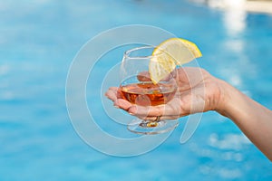 Glass of fragrant cognac with a slice of lemon in a woman`s hand against a background of blue pool water