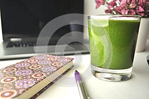 A glass of fresh green smoothie on a table with laptop computer, pink notebook, pen and a pink potted plant photo