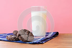 Glass of fresh milk with a pile of crunchy baked chocolate sugar cookies on wooden table with dark blue dinner napkin on pink back