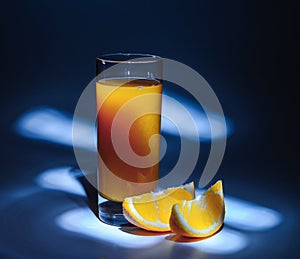 A glass of fresh orange juice and slices of orange on a blue background with backlight. In the dark tonality.