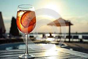 Glass of fresh summer cocktail on wooden table near swimming pool outdoors at sunset