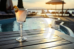 Glass of fresh summer cocktail on wooden table near swimming pool outdoors at sunset