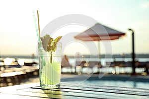 Glass of fresh summer cocktail on wooden table near swimming pool outdoors at sunset