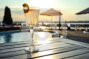 Glass of fresh summer cocktail on wooden table near swimming pool at sunset. Space for text