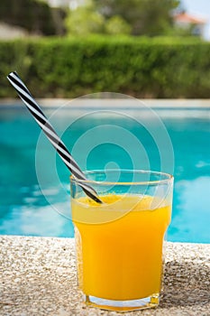 Glass of Freshly Pressed Tropical Fruits Orange Juice with Striped Straw on Deck of Villa Swimming Pool on Sunny Summer Day